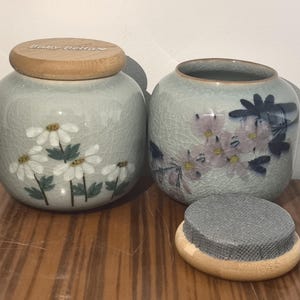 May include: Two ceramic jars with lids, one with a floral design and the other with a floral and dragonfly design. The jars have a light gray glaze and are decorated with white and pink flowers.