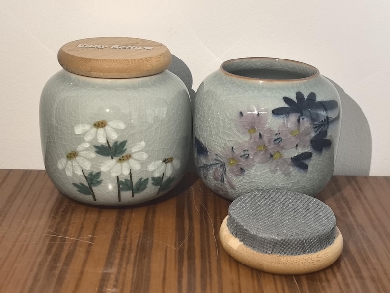 May include: Two ceramic jars with lids, one with a floral design and the other with a floral and dragonfly design. The jars have a light gray glaze and are decorated with white and pink flowers.