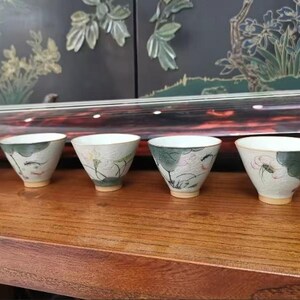 May include: Six small white ceramic teacups with green and pink floral designs. The cups are arranged in a row on a wooden surface.