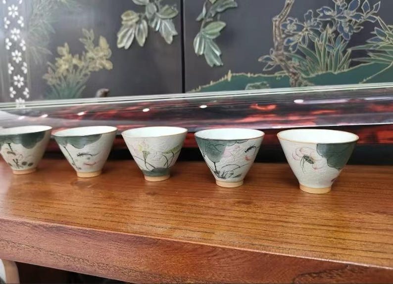 May include: Six small white ceramic teacups with green and pink floral designs. The cups are arranged in a row on a wooden surface.