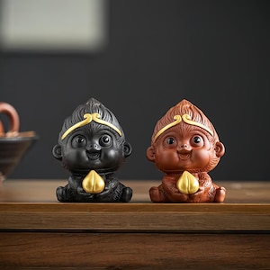 Può includere: Due piccole statuette di scimmie in ceramica, una nera e una marrone, entrambe con dettagli dorati e che tengono una pesca dorata nelle loro mani.