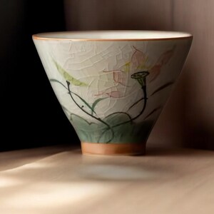 May include: A white ceramic teacup with a floral design. The cup has a wide, rounded base and a delicate rim. The floral design features pink and green flowers and leaves.