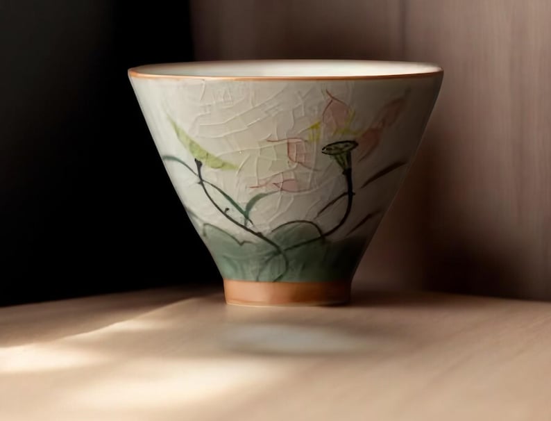 May include: A white ceramic teacup with a floral design. The cup has a wide, rounded base and a delicate rim. The floral design features pink and green flowers and leaves.