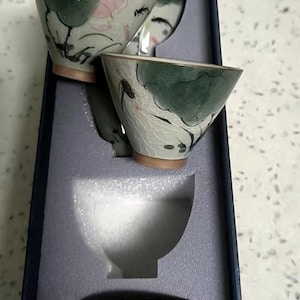 May include: Set of five small, white ceramic teacups with green and pink floral designs. The cups are stacked inside a blue box with white foam padding.