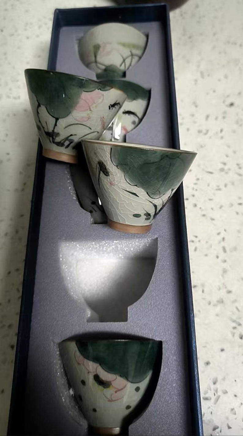May include: Set of five small, white ceramic teacups with green and pink floral designs. The cups are stacked inside a blue box with white foam padding.