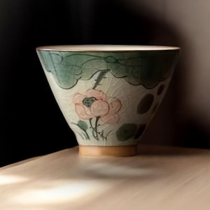 May include: A white ceramic teacup with a green and pink floral design. The cup has a wide, rounded base and a flared lip.