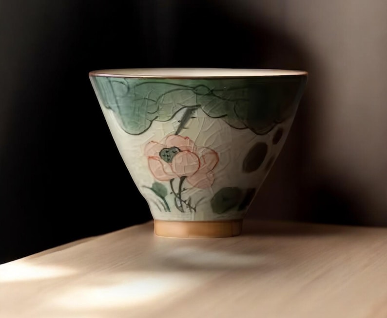 May include: A white ceramic teacup with a green and pink floral design. The cup has a wide, rounded base and a flared lip.