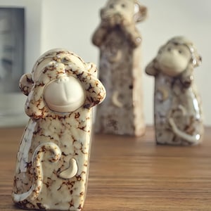 May include: Three ceramic monkey figurines, each with a different gesture: one covering its eyes, one covering its ears, and one covering its mouth. The monkeys are beige with brown marbling and have white faces.
