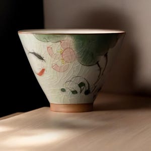 May include: A white ceramic teacup with a green, pink, and black floral design. The cup has a wide, rounded shape and a slightly flared rim.