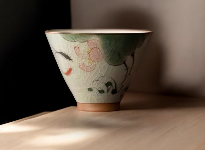 May include: A white ceramic teacup with a green, pink, and black floral design. The cup has a wide, rounded shape and a slightly flared rim.