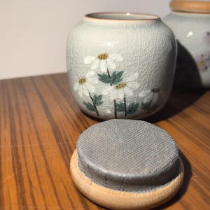 May include: Two ceramic jars with lids, one with a floral design of white flowers and green leaves on a light gray background, the other with a floral design of pink flowers on a light gray background.