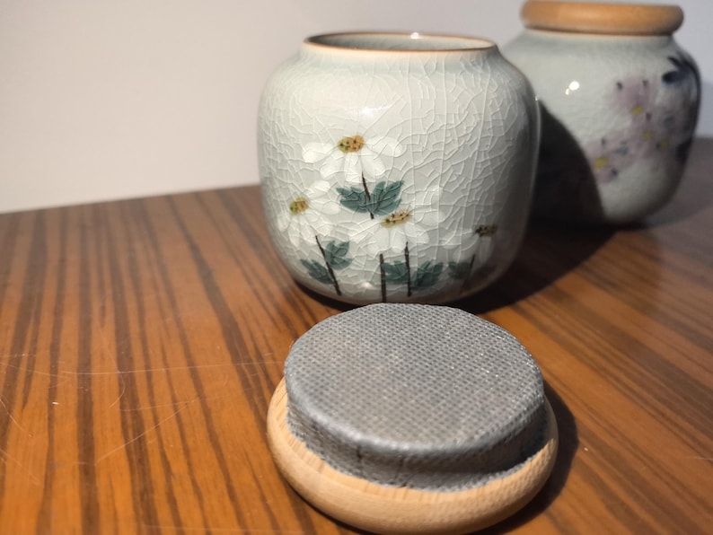 May include: Two ceramic jars with lids, one with a floral design of white flowers and green leaves on a light gray background, the other with a floral design of pink flowers on a light gray background.