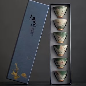 May include: A set of six small, green and white teacups with floral designs are displayed in a blue box. The box has the text "JINGKAI CHA SHI" printed on it.