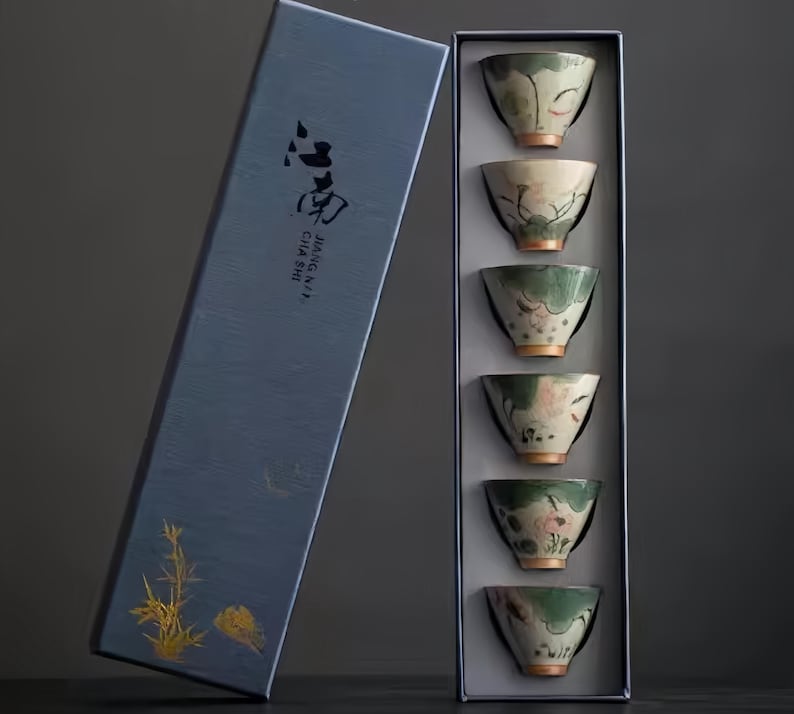May include: A set of six small, green and white teacups with floral designs are displayed in a blue box. The box has the text "JINGKAI CHA SHI" printed on it.