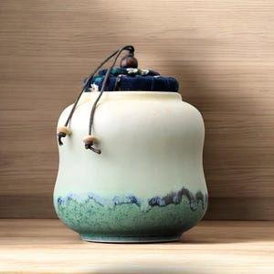 May include: A white ceramic jar with a green and blue glaze. The jar has a fabric lid with a floral pattern and a brown cord with wooden beads.
