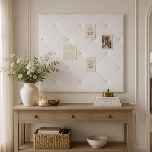 May include: A large, rectangular, white fabric-covered tufted bulletin board with diamond-shaped stitching and small buttons. The board is mounted on a wall and displays four small paper cards pinned to its surface in a grid-like pattern.
