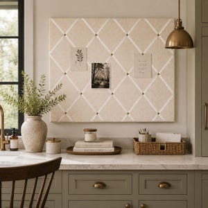May include: A rectangular beige fabric memo board with a white crisscross ribbon pattern secured by brass studs. The board displays three small paper prints and is mounted on a wall above a kitchen counter with a vase, books, and a basket.