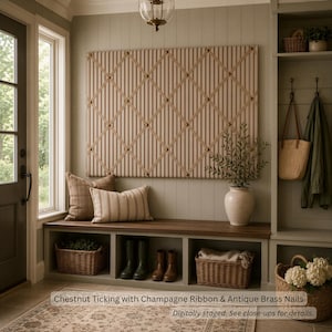 May include: A rectangular fabric memo board featuring chestnut ticking stripes, a champagne-colored ribbon lattice, and antique brass nail heads. The board is mounted on a wall above a wooden bench with storage cubbies and woven baskets.