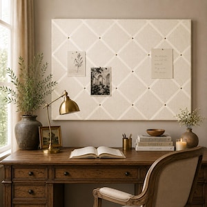 May include: A rectangular beige fabric memo board with a white ribbon diamond pattern secured by small brass studs. The board displays two paper cards, one with a botanical illustration and one with text that reads, The details are not the details. They make the design.