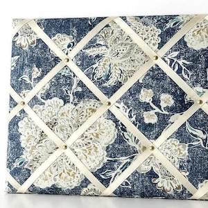 Puede incluir: Un tablero de notas rectangular cubierto de tela con un fondo azul oscuro de textura desgastada y un patrón floral blanco. El tablero está cruzado por cintas de color crema aseguradas con pequeños tachones metálicos dorados en cada intersección.