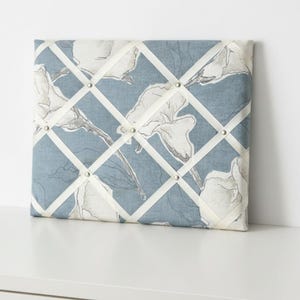 Puede incluir: Un tablero de notas rectangular de tela con un fondo azul polvoriento y un patrón de líneas florales blancas. El tablero está cruzado por cintas blancas aseguradas con pequeños tachuelas de metal plateado en cada intersección.