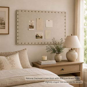 May include: A rectangular beige linen bulletin board featuring a border of antique brass nailhead trim. The board is mounted on a wall and displays four small rectangular cards pinned to its surface with brass-colored push pins.