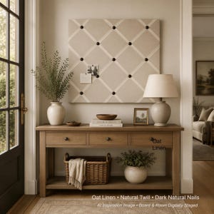 May include: A square beige fabric memo board with a white ribbon grid and dark nail head accents. The board is mounted on a wall above a wooden console table featuring three drawers, a woven basket, and a potted plant on the lower shelf.