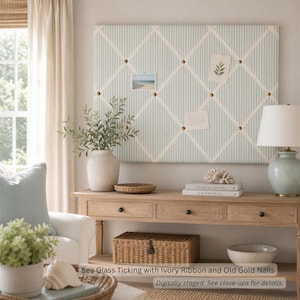 Puede incluir: Un tablero de notas rectangular con rayas tipo ticking en azul claro y blanco, cruzado con cintas de color marfil aseguradas por clavos dorados. El tablero muestra dos pequeñas notas de papel y está montado sobre una consola de madera.