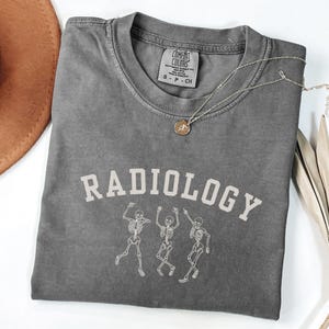 Puede incluir: Una camiseta gris oscura con el texto "RADIOLOGY" en letras blancas. Tres esqueletos blancos bailan debajo del texto. La camiseta tiene un aspecto desgastado.