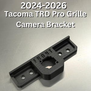 May include: A black camera bracket for a Tacoma TRD Pro Grille. The bracket has the word "FRONT" printed on it and is designed for the 2024-2026 model years. It features a rectangular design with mounting holes.