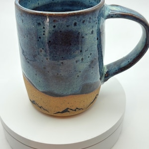 May include: A handmade ceramic mug with a blue glaze and a tan base. The mug features a mountain range design around the base. The mug has a curved handle and is suitable for hot or cold drinks.