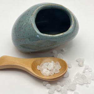May include: A smooth, rounded, blue-grey ceramic bowl with a speckled finish and a wide opening, accompanied by a light-coloured wooden spoon filled with coarse white salt crystals, with additional salt crystals scattered nearby.
