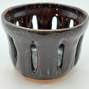 May include: A dark brown ceramic bowl with vertical openings around the sides. The bowl has a glossy finish and a slightly uneven texture. The interior is a darker shade of brown.