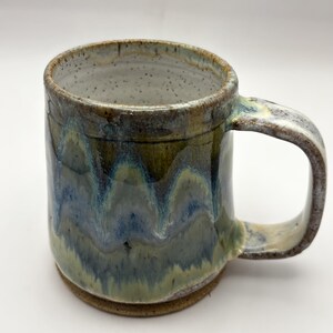 May include: A ceramic mug with a speckled white interior and a glossy exterior featuring a layered glaze of olive green, deep blue, and earthy brown tones. The mug has a sturdy, thick handle and a cylindrical body with a visible, unglazed brown base.