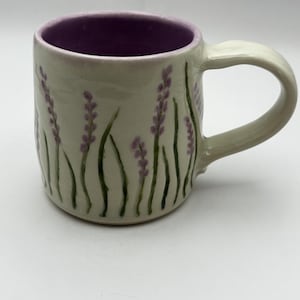 Peut inclure: Une tasse en céramique avec un extérieur blanc cassé brillant orné de brins de lavande violets et de tiges vertes peints à la main. L'intérieur de la tasse est d'un violet vif uni. Elle possède une anse incurvée robuste et un corps cylindrique.