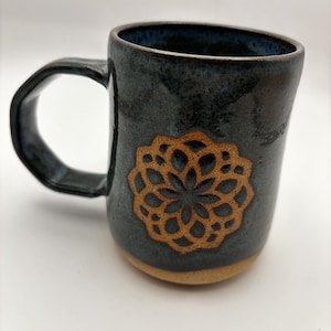 May include: A ceramic mug with a dark blue, glossy glaze and a textured, unglazed tan base. The mug features a prominent, embossed golden-brown mandala-style floral pattern on the front and a sturdy, angular handle extending from the side.