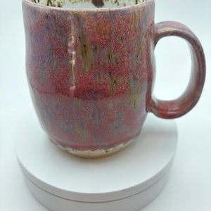 Peut inclure: Une tasse en céramique faite à la main avec un glaçage rouge et marron. La tasse a un corps arrondi, une grande anse et un bord moucheté. Le glaçage a une finition brillante. La tasse est posée sur une surface blanche.