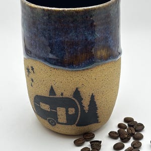 May include: A ceramic tumbler with a speckled tan base and a dark blue glazed rim. The exterior features a black graphic print of a vintage camper trailer surrounded by pine trees and small stars. The top edge has a small rectangular cutout.