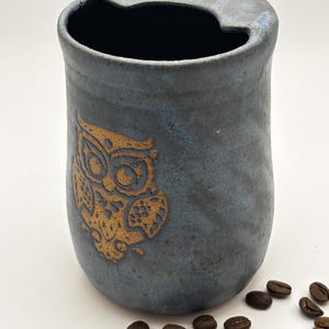 May include: A cylindrical ceramic tumbler with a matte blue-grey finish and a textured surface. The front features a stamped golden owl design. The rim has a small cutout notch and a single hole, resembling a travel cup lid design.