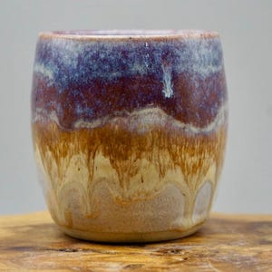 Peut inclure: Une tasse en céramique avec un glaçage brun, jaune, violet et blanc. La tasse a une forme arrondie et un bord légèrement évasé.