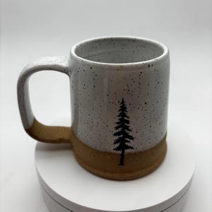 May include: A speckled white ceramic mug with a brown base and handle. A black pine tree design is on the front of the mug. The mug is on a white surface.