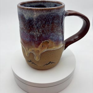May include: A handmade ceramic mug with a dark brown handle and a body that transitions from a deep purple to a tan base. The base features a mountain range design. The mug is approximately 10 cm tall.