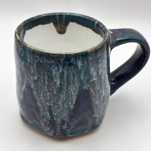 May include: A ceramic mug with a glossy glaze featuring a blend of deep blue, purple, and white colours that create a dripping, textured effect. The interior of the mug is a solid, smooth white, and it includes a sturdy, curved handle on the side.