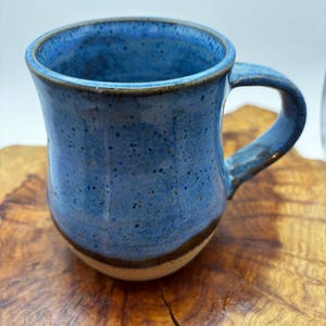 May include: A blue ceramic mug with a speckled glaze. The mug has a curved handle and a brown band near the base. The mug is displayed on a wooden surface. The mug is suitable for hot beverages.