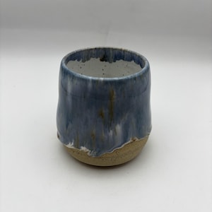 May include: A ceramic cup with a rounded shape, featuring a smooth, glossy blue and white drip glaze on the upper portion and a textured, speckled beige clay base. The interior is white with small dark speckles throughout the surface.
