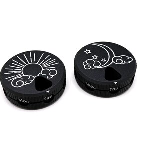 May include: Two black, round pill organizers with white graphics. One features a sun and clouds, the other a moon, stars, and clouds. Each has a rotating dial with day labels, such as "Mon" and "Tue".