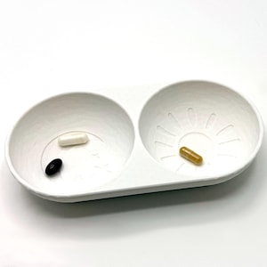 Op de afbeelding: Een witte pillendoos met twee compartimenten. Eén compartiment bevat een zwarte pil en een witte pil, terwijl het andere compartiment een bruine pil bevat.