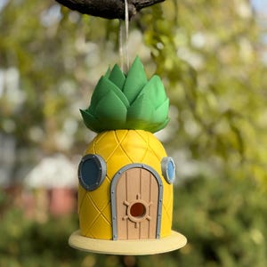 May include: A yellow pineapple-shaped birdhouse with a green leafy top, a brown wooden door, and two blue porthole windows. The birdhouse is hanging from a branch.