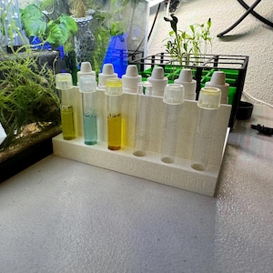 May include: A white plastic test tube rack with 10 clear glass test tubes. The test tubes are filled with different colored liquids, including blue, yellow, and clear.