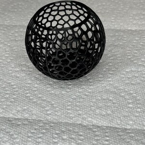 May include: A black, spherical object with a honeycomb-like pattern. The intricate design features a series of interconnected hexagonal shapes, creating an open, lattice-like structure. The object is likely a decorative item or a component of a larger design.